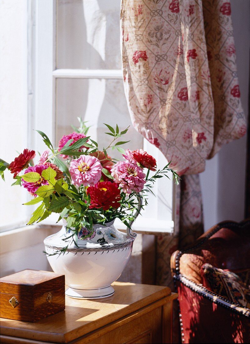 Ceramic vase of garden flowers in sunlight in front of open lattice window with draped curtain