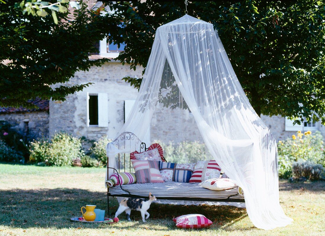 Eisenbett mit gemusterten, handgefertigten Kissen unter drapiertem Moskitonetz in einem französischen Landhausgarten