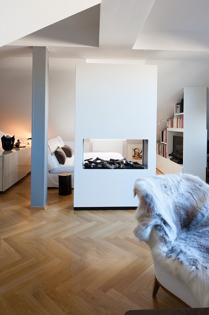 Converted attic with herringbone parquet floor, fireplace in partition between sleeping area and living area with fur blanket on armchair