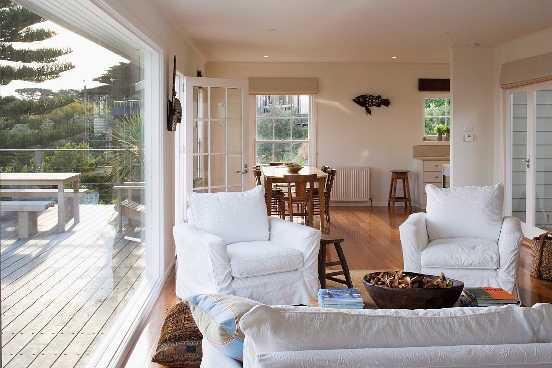 Offener Wohnraum im Landhaus - Sessel mit weisser Husse um Couchtisch und Essplatz im Hintergrund, an der Seite Glasfront mit Blick auf möblierte Holzterrasse