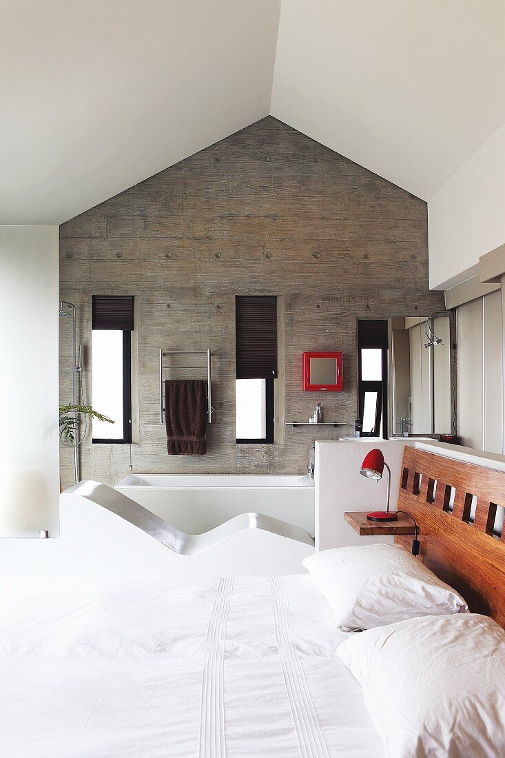 Blick von Doppelbett mit integriertem Kopfteil auf die Betongiebelwand der Bad Ensuite; Einbau-Liege als Raumteiler