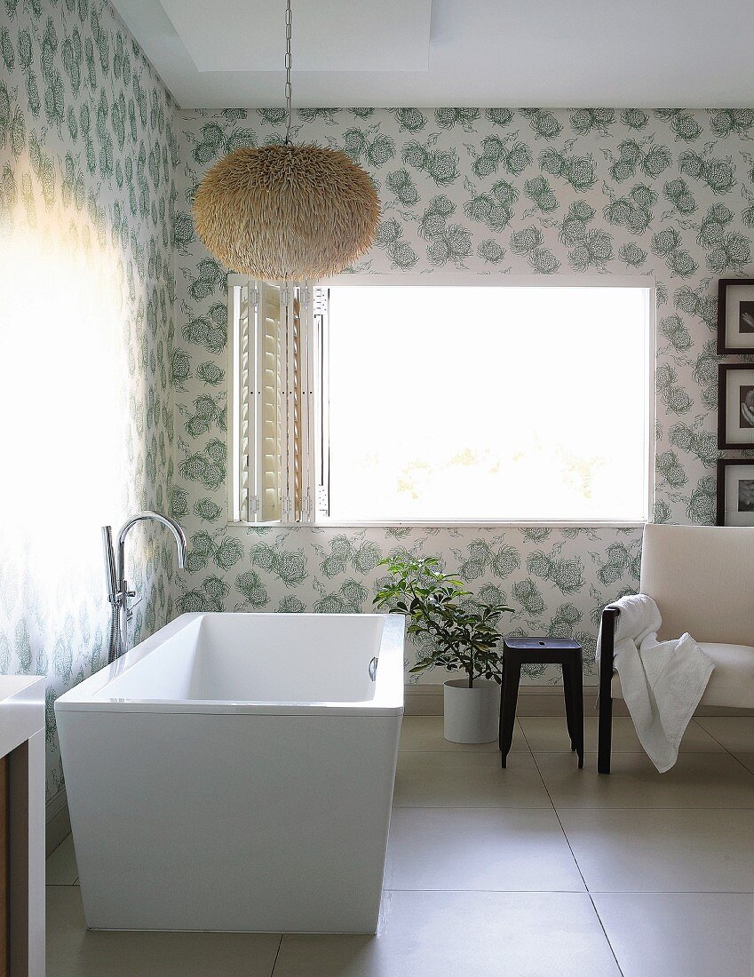 Moderne freistehende Badewanne mit Standarmatur unter puscheliger Hängeleuchte in Badezimmerecke mit floralem Muster auf tapezierter Wand, im Hintergrund offenes Fenster mit faltbaren Fensterläden