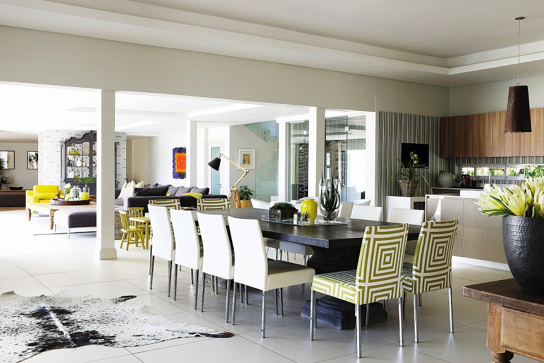 Gepolsterte Stühle teilweise mit gemustertem Bezug an langer Esstafel auf weißem Fliesenboden, Loungebereich hinter Stützen in offenem Wohnraum