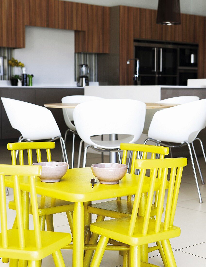 Gelber Kindertisch mit Stühlen vor modernen, weissen Schalenstühlen in offener Küche