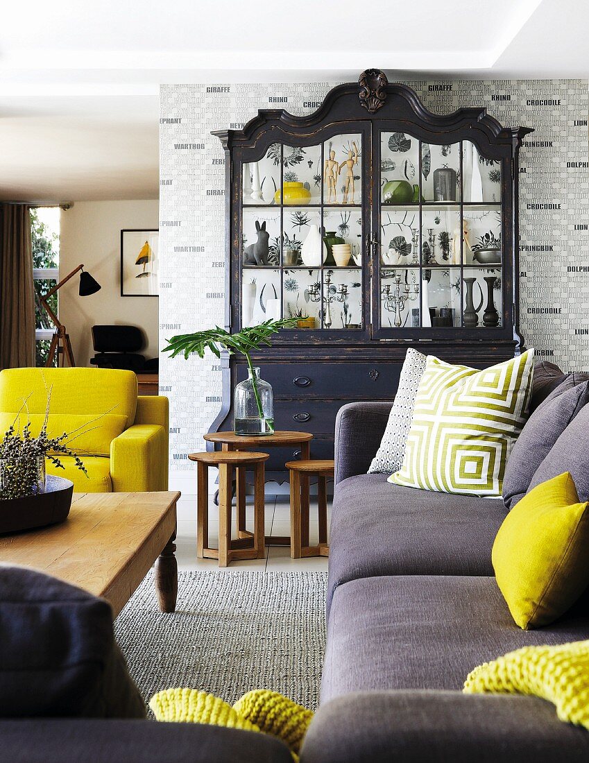 Sofa with bright yellow and patterned scatter cushions in front of rustic wooden coffee table, black-painted dresser with glass doors against patterned wallpaper