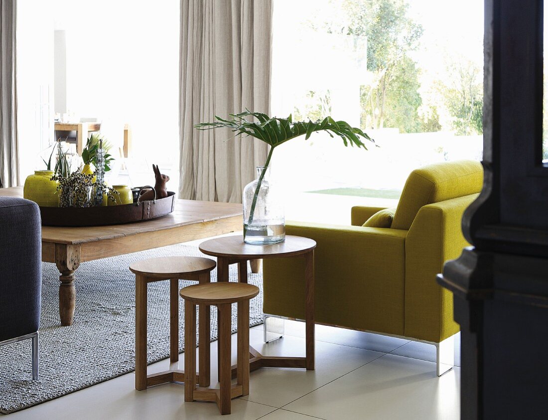 Mehrteiliges Beistelltischset aus Holz, neben gelbem Sessel gegenüber rustikalem Holz Couchtisch mit Tablett und verschiedenen gelben Vasen, im Hintergrund Fensterfront mit Gartenblick