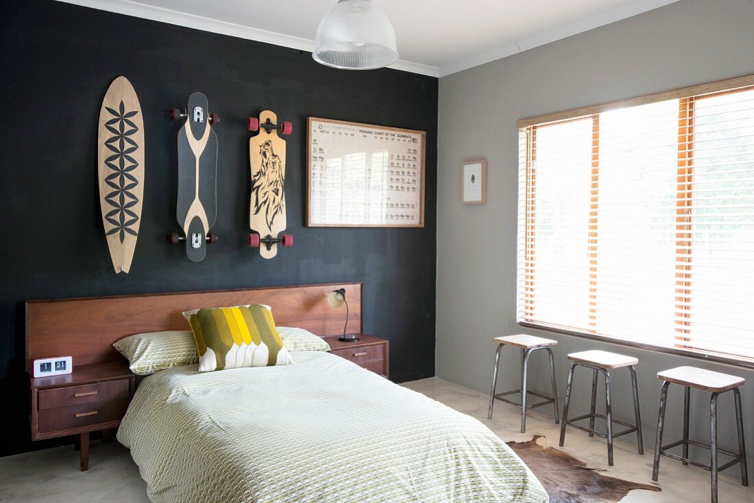 Bett mit Holz Kopfteil vor schwarz getönter Wand, daran aufgehängte, bemalte Skateboards; an der Seite drei Hocker vor Fenster mit geschlossener Jalousie
