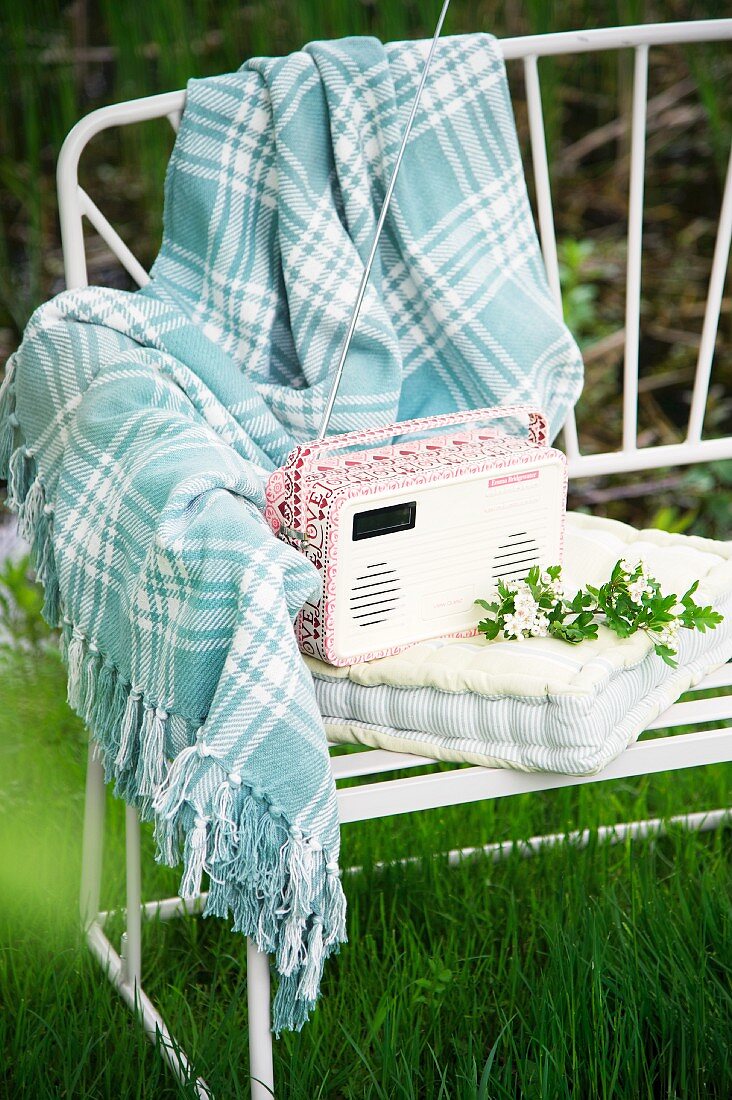 Kleines Kofferradio auf Polsterkissen und karierte Wolldecke auf filigraner Gartenbank aus weißem Metall im Freien