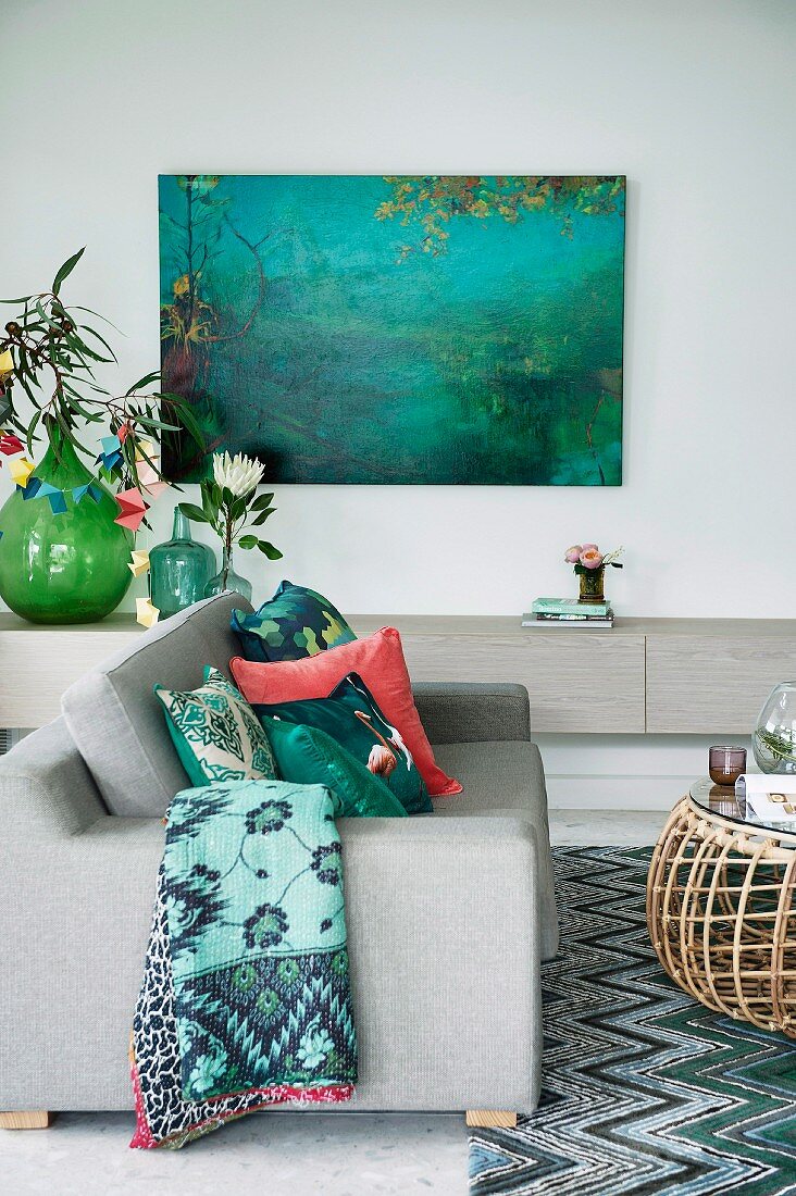 Sofa und Sideboard in Hellgrau, harmonisch mit grünen Accessoires wie Textilien, Glasballon und einem Gemälde kombiniert