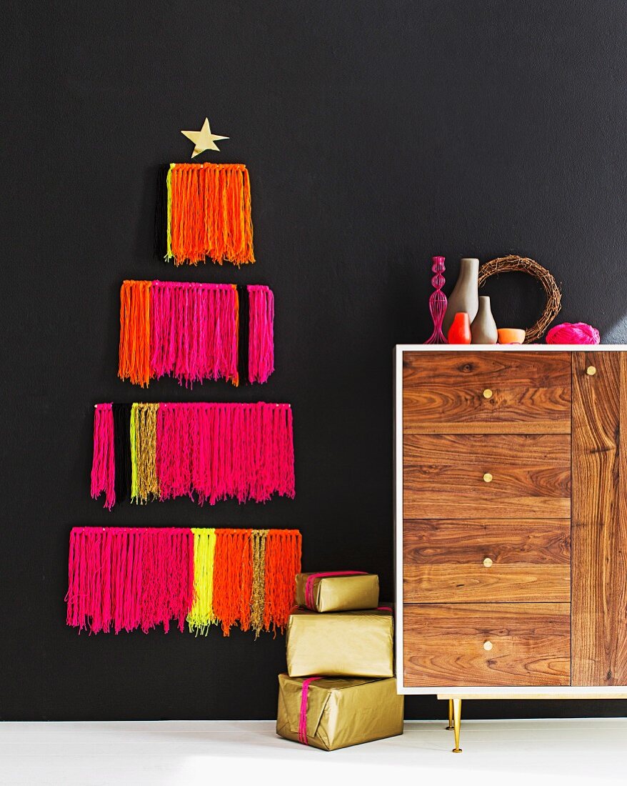 Stylised Christmas tree made from neon woollen fringes on black wall, arrangement of vases on retro sideboard and stacked presents on floor