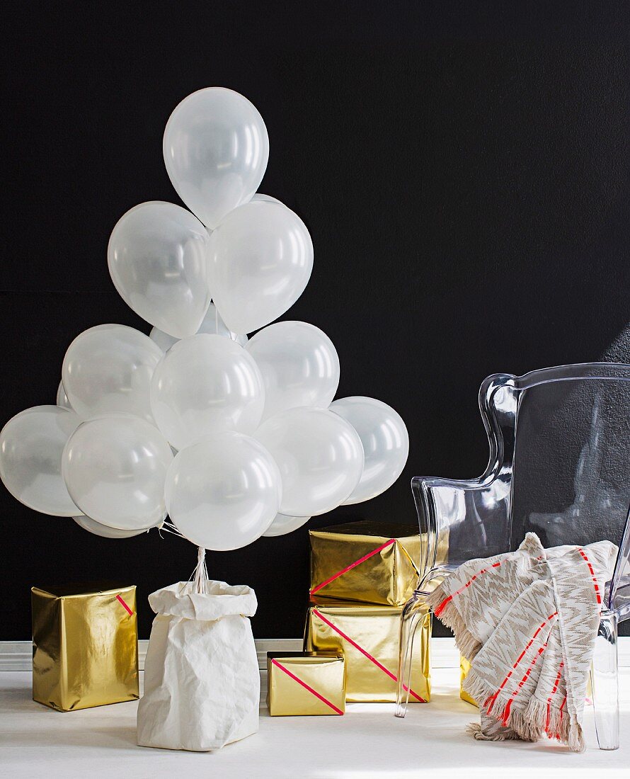 Mit weißen Luftballons stiliesierter Weihnachtsbaum und Geschenke in Goldpapier vor schwarzem Hintergrund