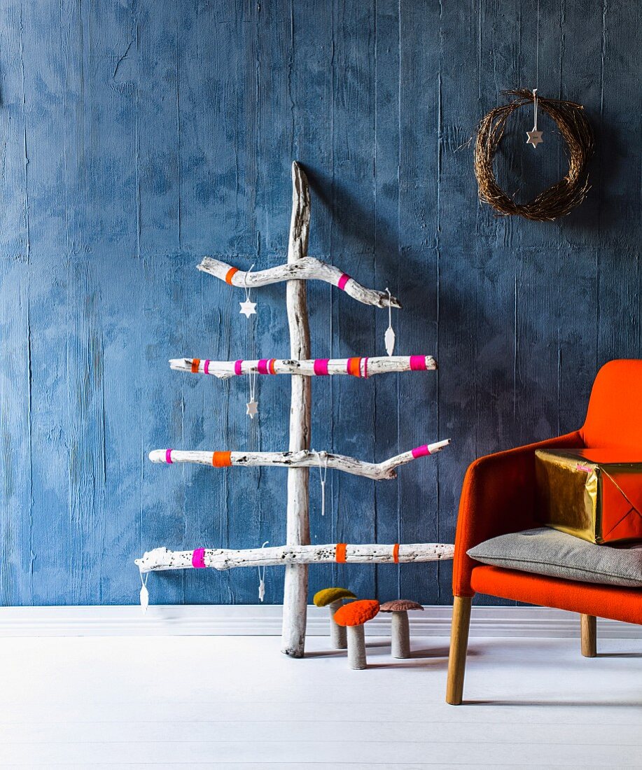 Stilisierter Weihnachtsbaum aus Strandgut gezimmert mit Weihnachtsanhängern geschmückt an blauer Wand und Dekopilzen am Boden