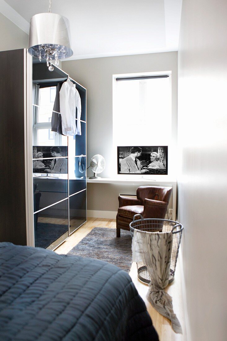 Kleiderschrank mit Schiebetüren aus schwarzem Glas, brauner Ledersessel vor halbhohem Regal mit Fernseher am Fenster im Schlafzimmer