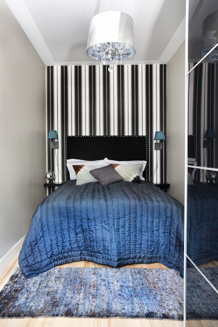 Gesteppte Tagesdecke auf Doppelbett mit schwarzem Kopfteil in schmaler Nische, vor schwarzweisser Streifentapete an Wand, am Bettende Teppichläufer auf Holzboden