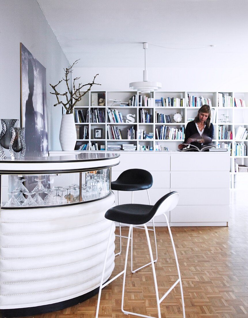 Bar stools at semicircular bar with glass top, woman in background next to half-height, white chest of drawers and bookcase