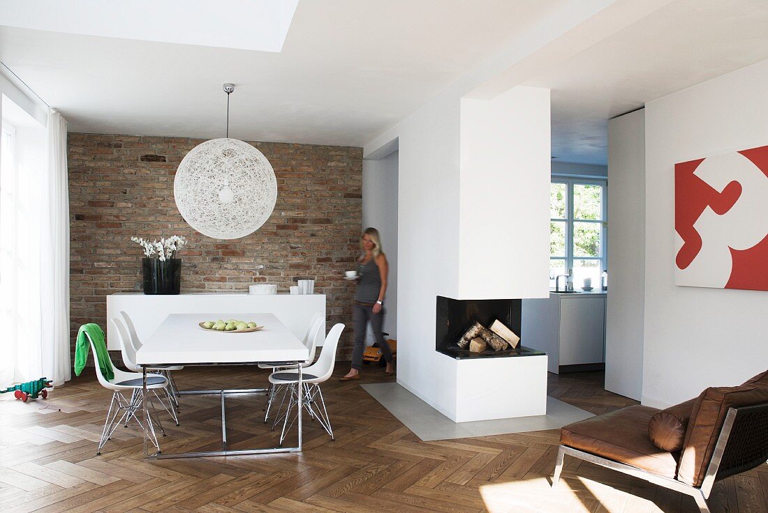 Open-plan interior with dining area, free-standing fireplace block and herringbone parquet floor; exposed brick wall behind dining area
