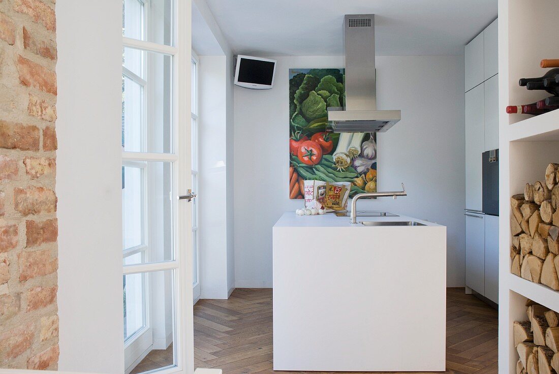 Blick in kleine, weiße Designerküche mit zentralem Arbeitsblock; Gemüsebild und Wandmonitor im Hintergrund
