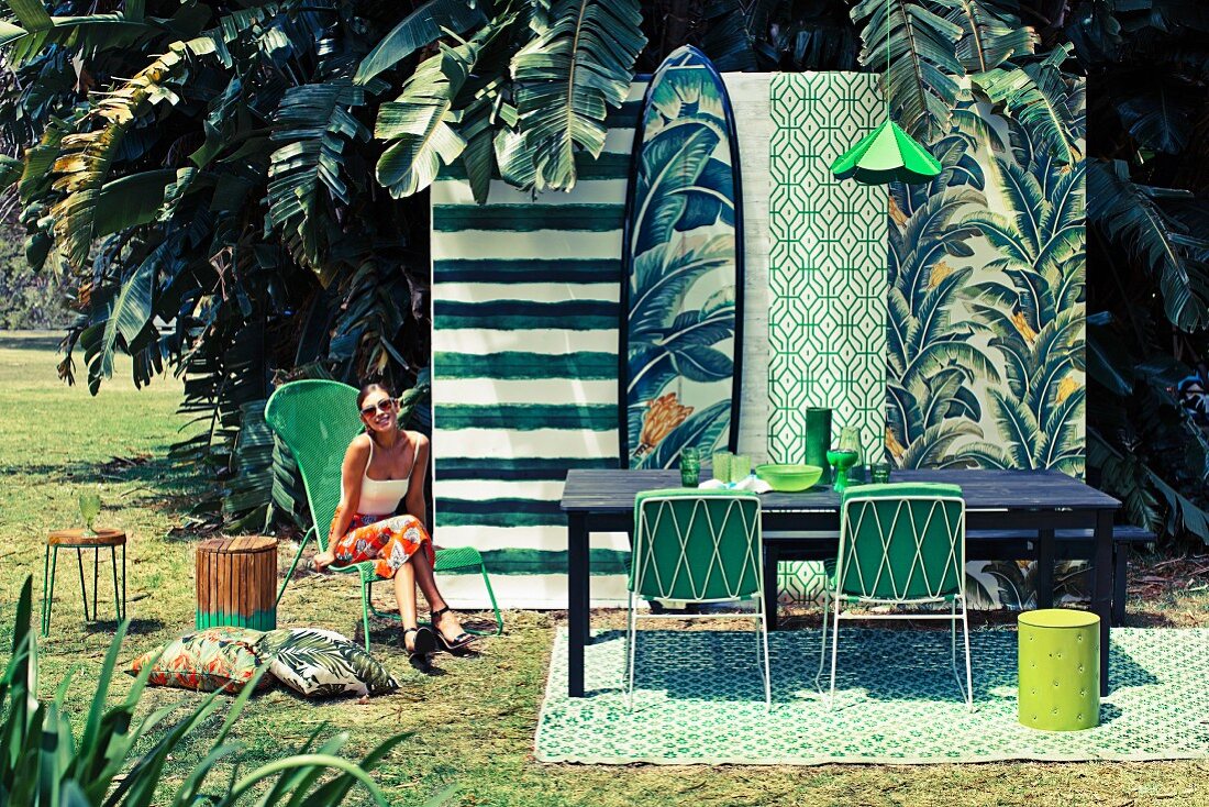 Tapetenstreifen mit grünem Mustermix als Hintergrundkulisse und mit passenden Accessoires dekorierter Essplatz im Freien; junge Frau auf einem Stuhl