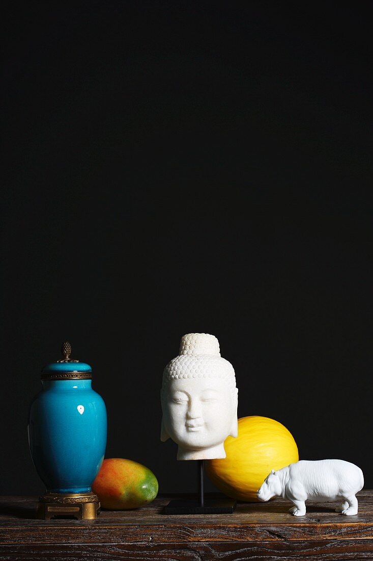 Arrangement mit weißem Buddhakopf und Nilpferd-Figur, Früchten und blau glasierter Urnenvase vor dunklem Hintergrund