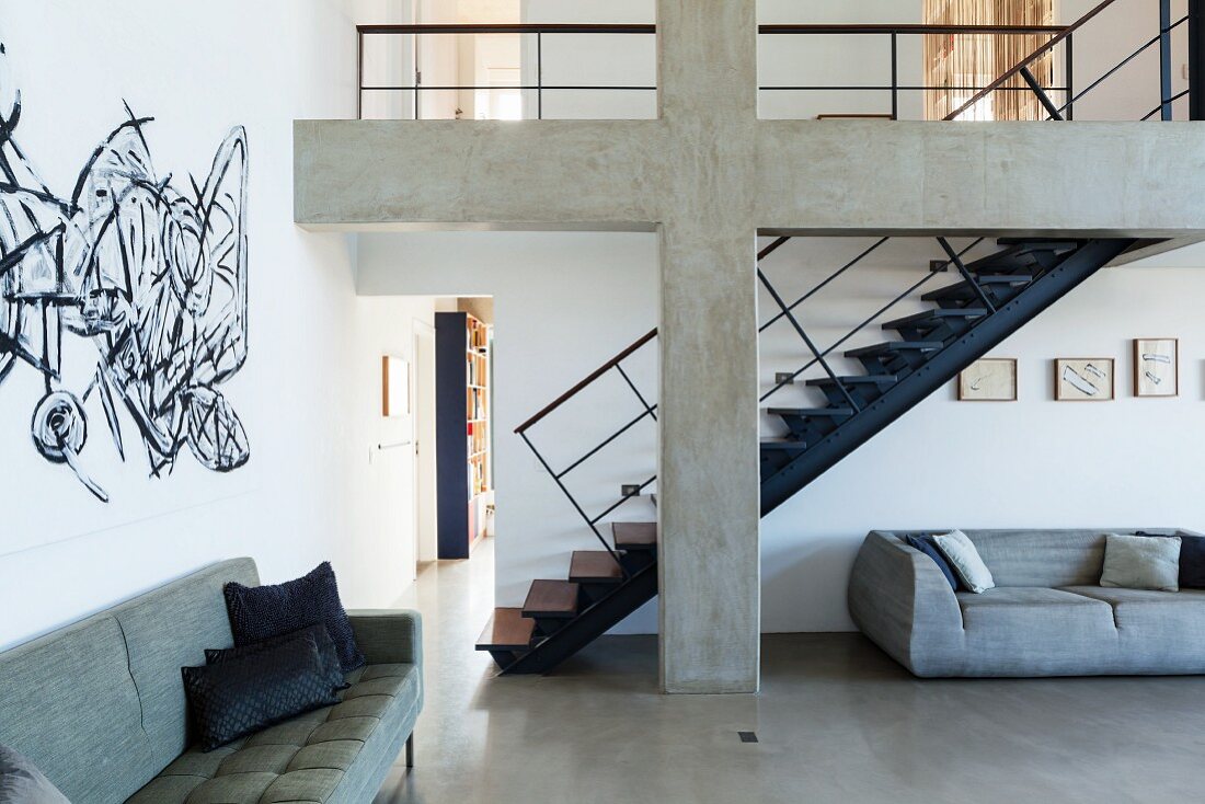 Dunkelblaue Stahltreppe hinter einem Betonkreuz in offenem Loft Wohnraum mit grauen Sofas und Wandmalerei im Graffiti Stil