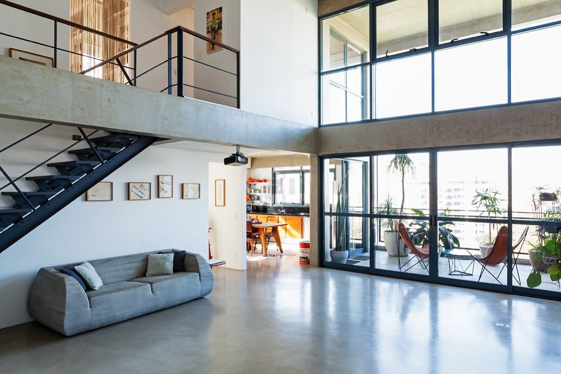 Offener Wohnraum in Betonskelettbau mit Stahltreppe und poliertem Betonboden; überdachte Terrasse hinter zweigeschossiger Glasfassade
