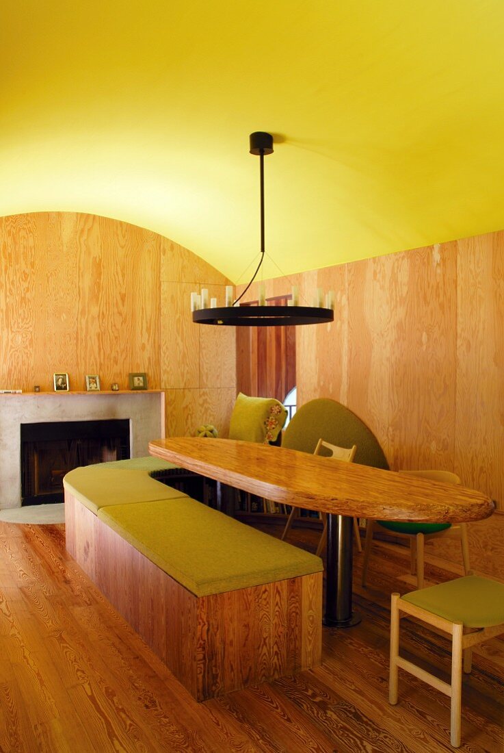 Vaulted ceiling painted lime green in interior with dining table and curved, wooden bench in front of modern, open fireplace