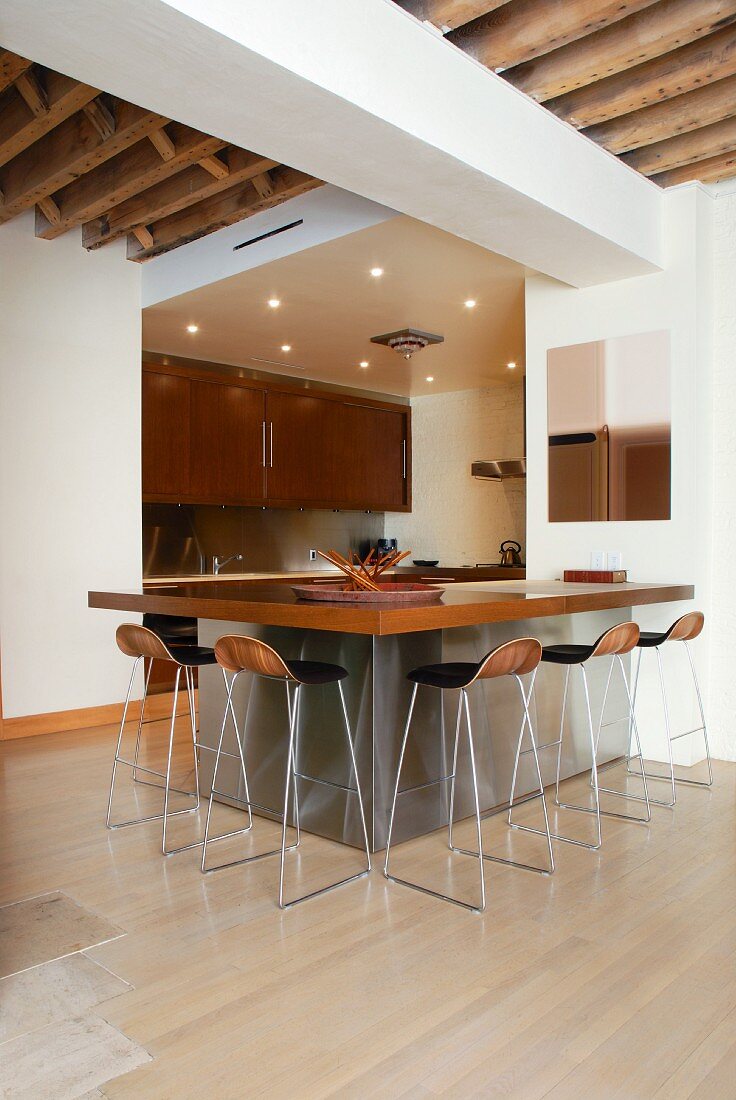Designer bar stools at kitchen counter with wooden worksurface on stainless steel base in open-plan interior