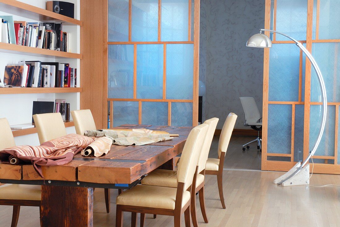 Chairs with pale upholstery at rustic wooden table with bolts of cloth and arc lamp in front of wooden-framed sliding wall with Japanese paper panels