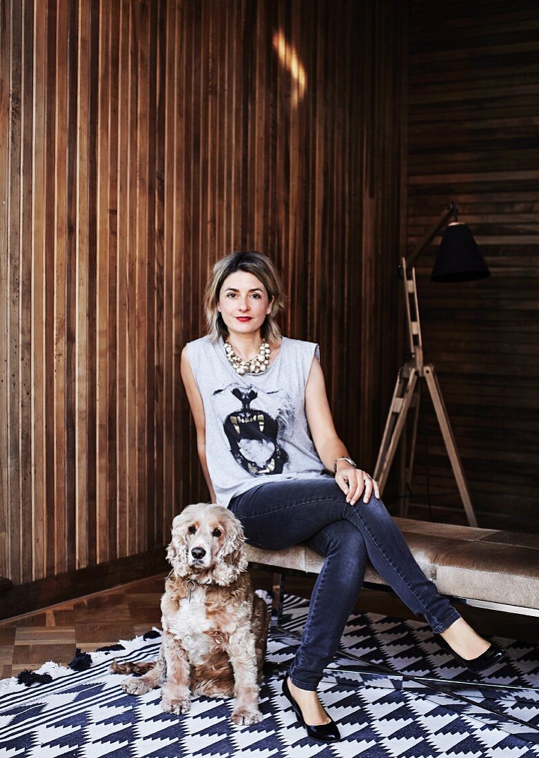 Frau auf Sitzbank und Hund auf Teppich mit geometrischem Muster vor Holzwand