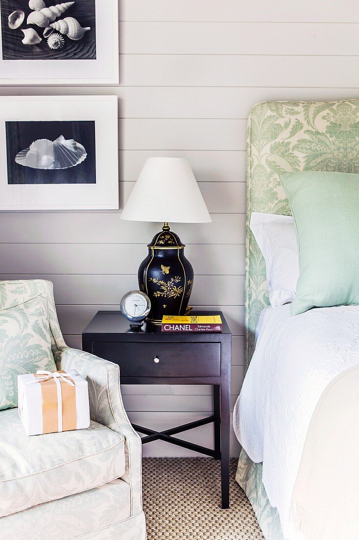 Black bedside cabinet with Chinese table lamp between armchair and bed against pale grey wooden wall