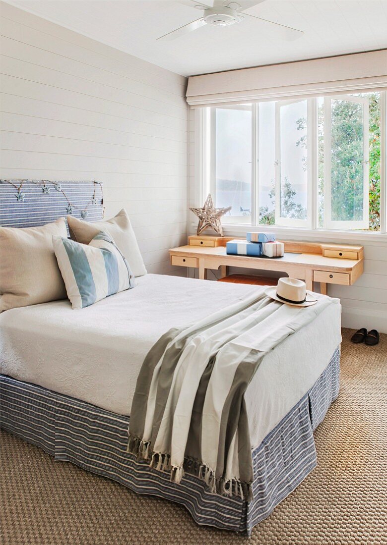 Kissen auf Doppelbett mit Kopfteil an hellgrauer Holzwand neben Schreibtisch aus Holz am Fenster in schlichtem Schlafzimmer
