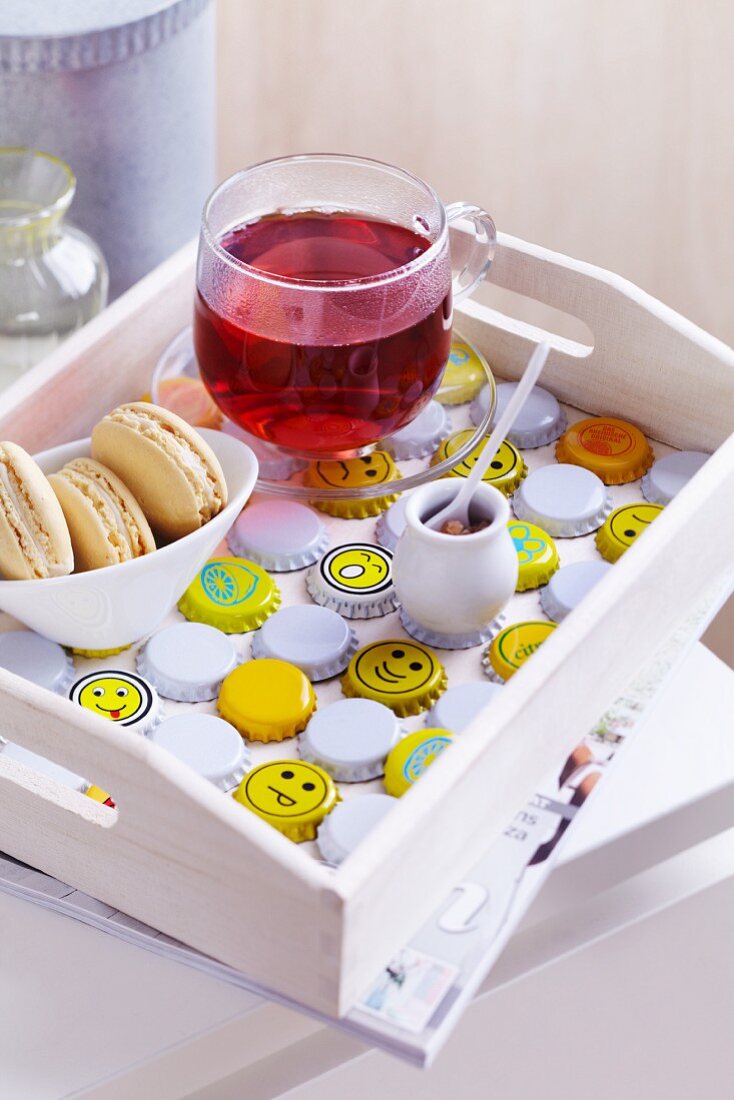 White wooden tray with cup of fruit tea and dish of biscuits arranged on bottle caps painted white and yellow with smiley faces
