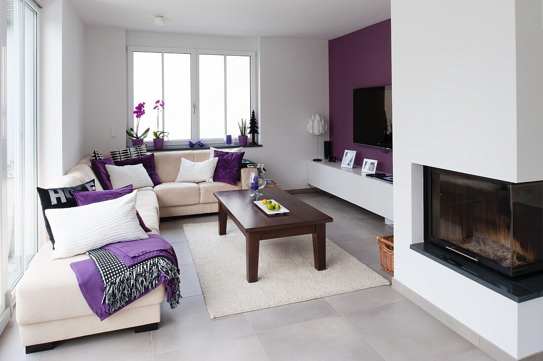 Heller moderner Wohnraum mit weißem Überecksofa um dunklen Holz Couchtisch und weißem Teppich, gegenüber violett getönte Wand mit offenem Kamin