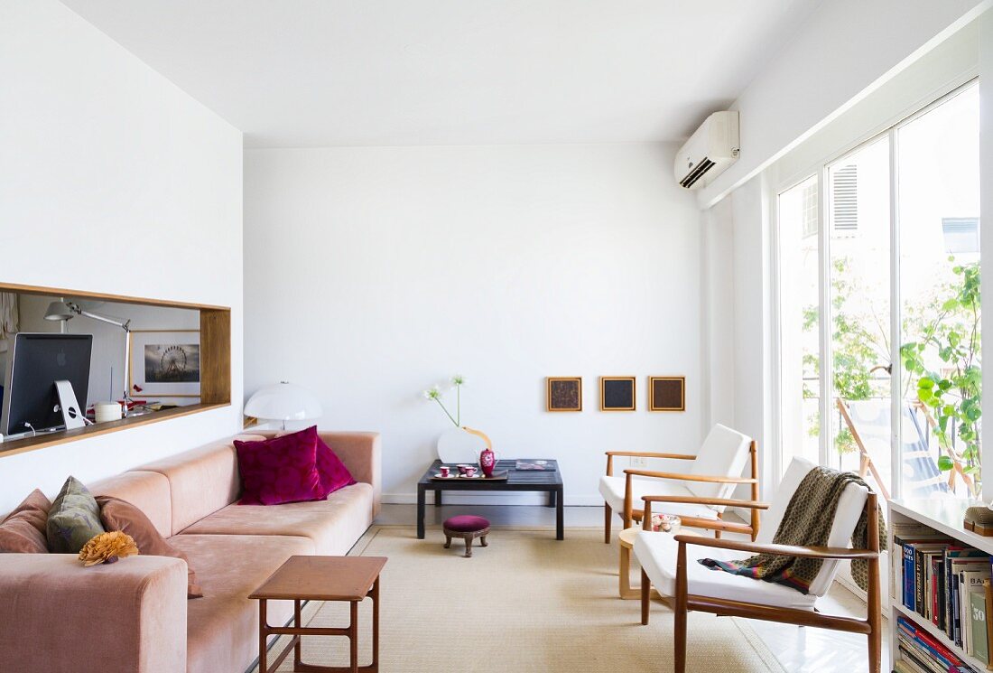 Minimalistischer, weisser Wohnraum - Pastellrosa Polstercouch und Beistelltisch gegenüber Sessel vor Fensterfront, Wandausschnitt mit Blick ins Arbeitszimmer