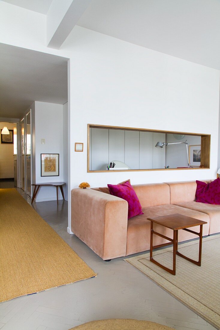 Offener Wohnraum mit Loungebereich, pastellrosa Couch und Holz Beistelltisch vor Wand mit fensterartiger Öffnung, seitlich Blick in beleuchteten Gangbereich
