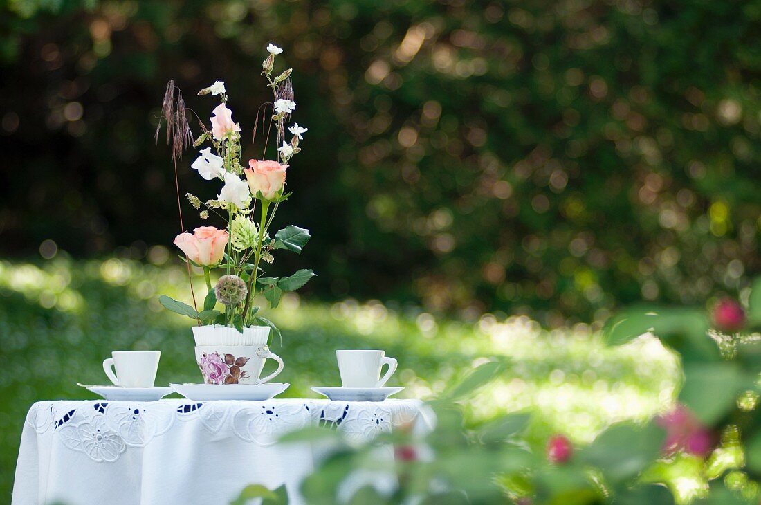 Romantisches Rosengesteck in nostalgischer Tasse auf Gartentisch mit weißer Tischdeckeund zwei Kaffeetassen