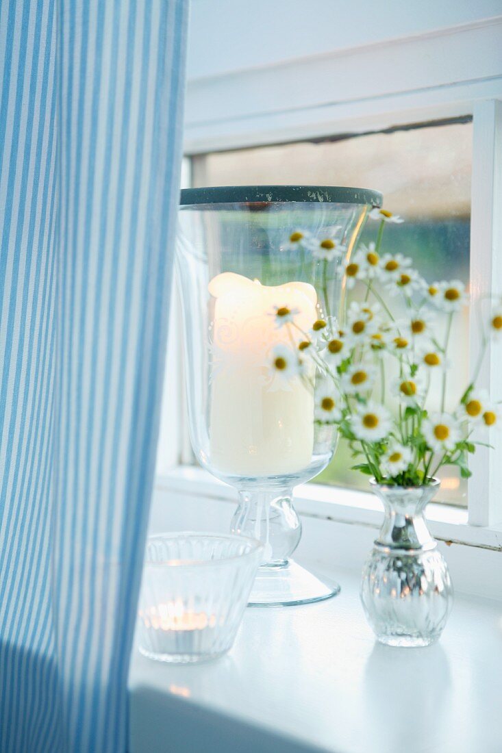 Windlicht und Teelicht mit Kamillenstäußchen auf weißem Fenstersims mit hellblau gestreiftem Vorhang