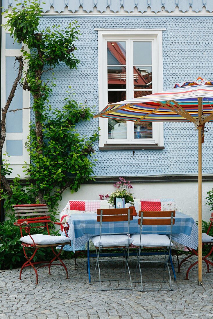 Lauschiger Terrassenplatz mit Sonnenschirm und Gartenstühlen vor hellblauem Schindelfassaden-Landhaus