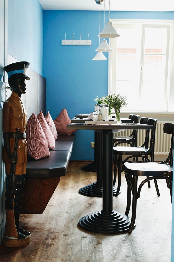 Gastraum mit Bistrotischen und Holzstizbank mit Kissen, blauen Wänden und maskuliner Holzfigur an der offenen Tür