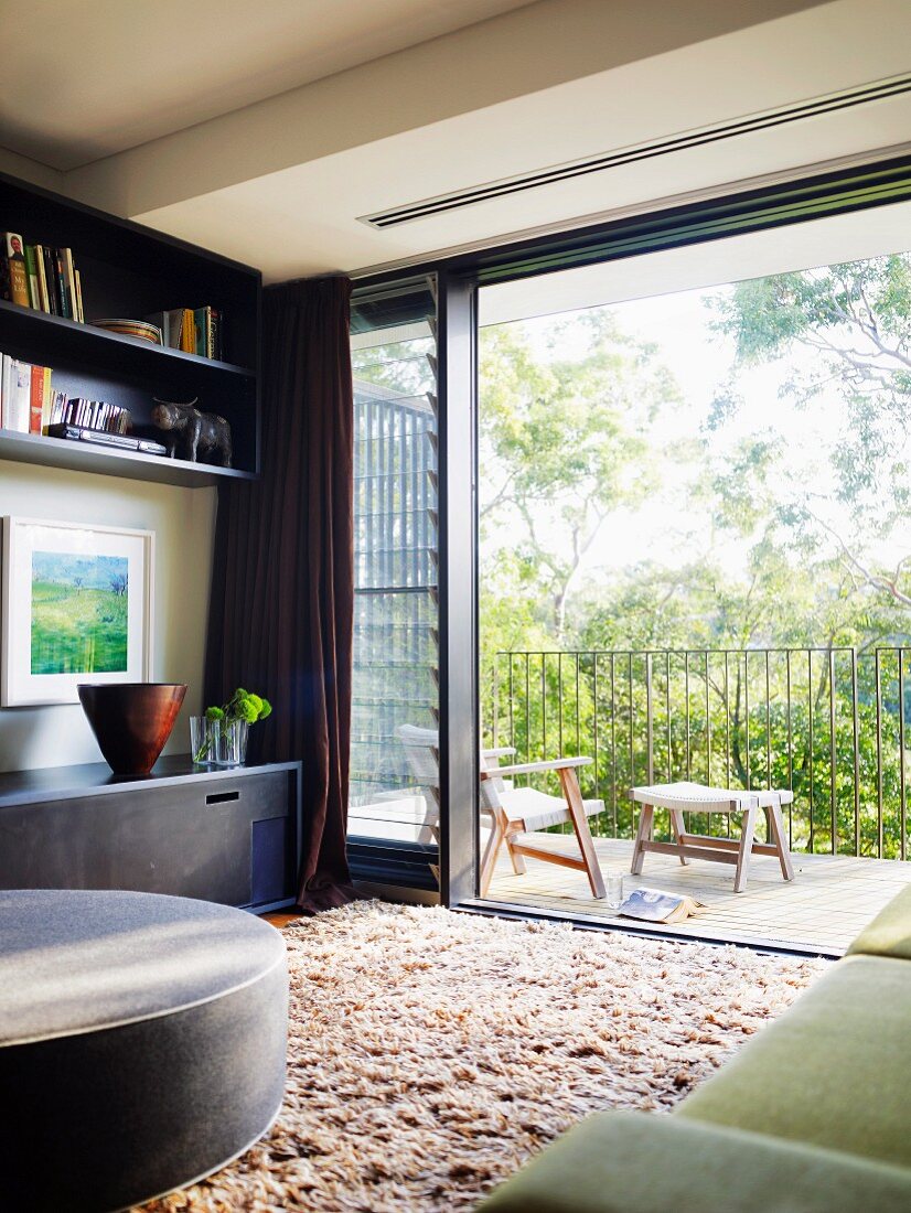 Blick von Wohnzimmer mit Hochflorteppich durch weit geöffnete Glasschiebetür auf Balkon