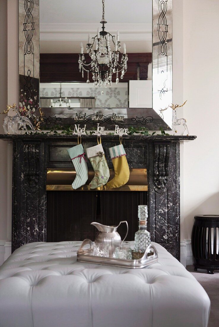 Fireplace decorated with Christmas stocking, garlands and glass reindeer below elegant mirror; silver tray on ottoman in foreground