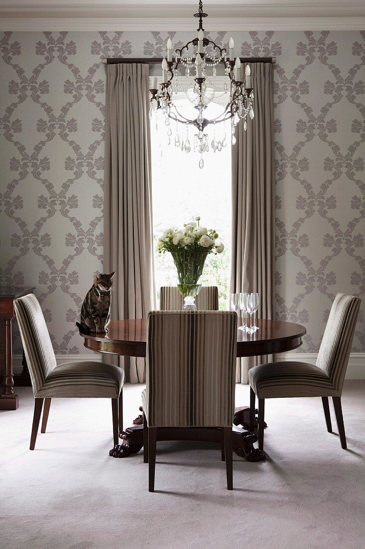 Chandelier above Georgian-style round table, striped upholstered chairs and floral wallpaper in elegant dining area in subtle shades of grey