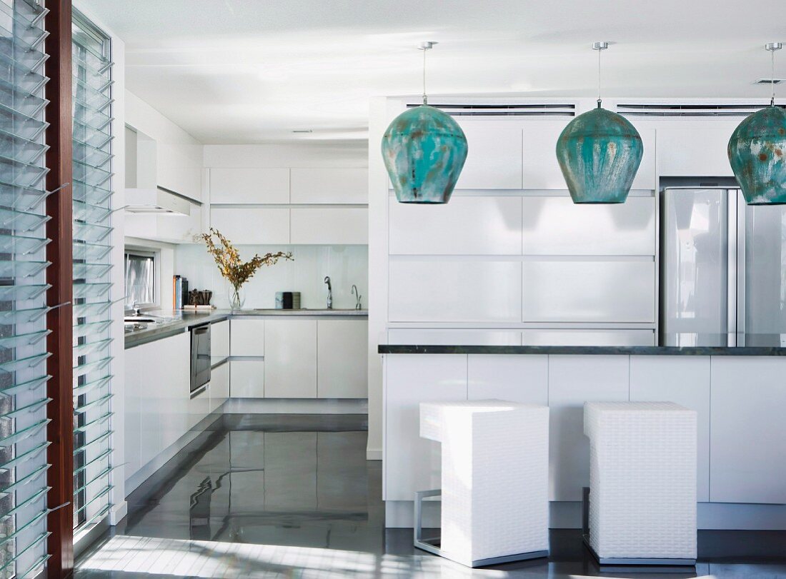 Moderne, offene Küche in Weiß, über Theke Hängeleuchten mit bauchigem Schirm aus Metall, vor der Theke weiße, kubische Barhocker im Designer Stil
