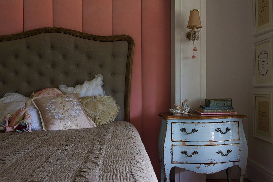 Bett mit romantischen Kissen und gepolstertem Kopfteil vor altrosa Wandverkleidung, daneben antike hellblaue Kommode mit Patina