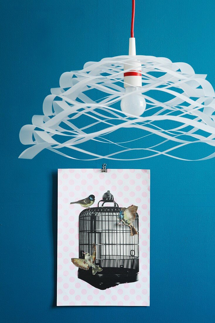 Luftige Papier-Pendelleuchte vor blauer Wand mit Collage aus ausgeschnittenen Papiervögeln am Vogelkäfig