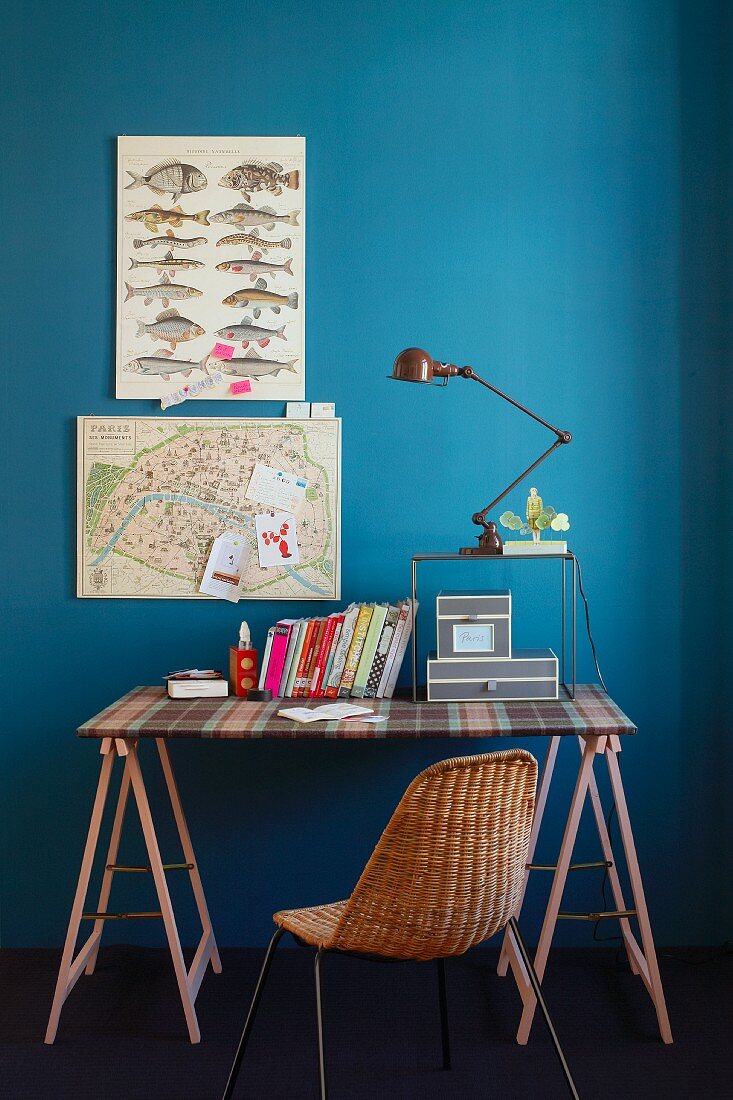 Desk with top covered in tartan fabric and wooden trestles, grey cardboard boxes, books, desk lamp and old map