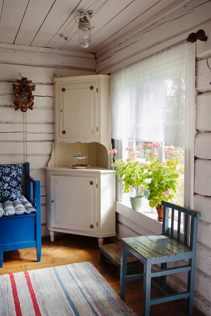 Corner of rustic living room in wooden house - white corner cupboard next to window and blue kitchen chair