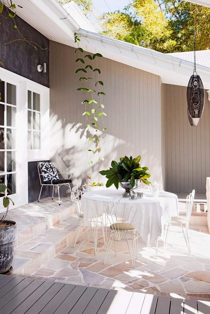 Sonnenbeschienene Terrasse mit filigranen Metallstühlen und Tisch auf Natursteinboden vor Holzhaus