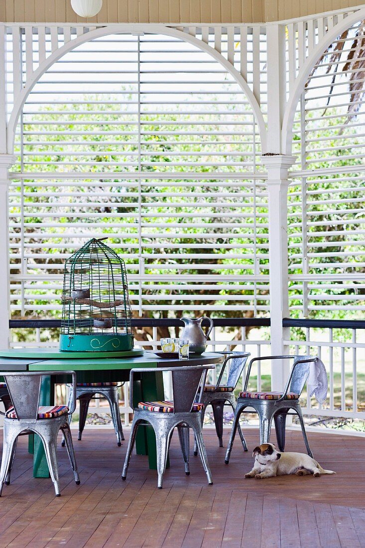 Birdcage on table and retro-style metal chairs in garden pavilion with slatted screens