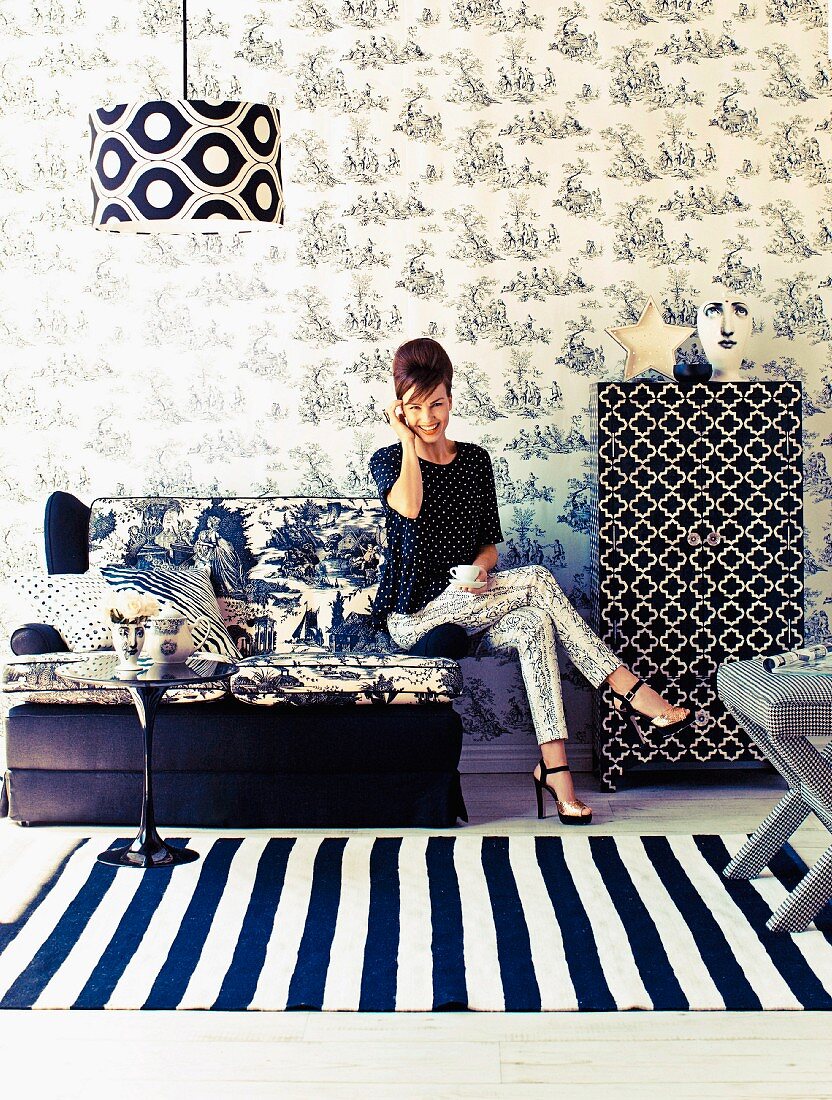 Seating area in fashionable mixture of black and white patterns; woman seated on two-seater sofa, floral wallpaper and striped rug